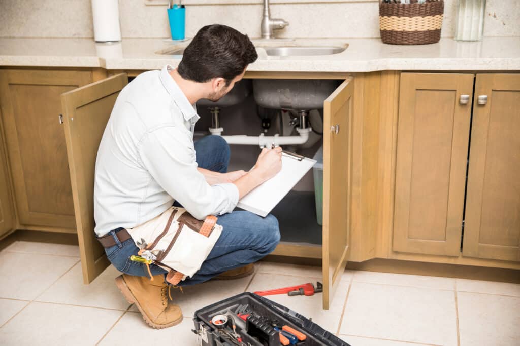 Plumber crouching by kitchen sink writing on a clipboard